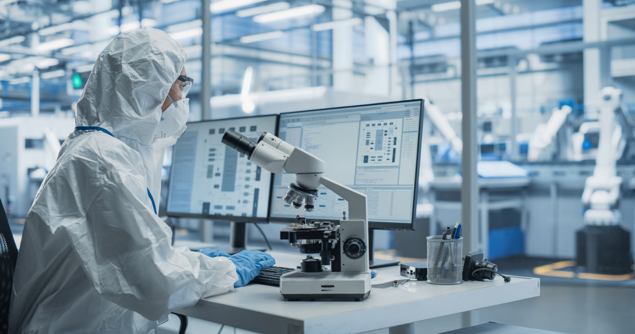 Research Factory Cleanroom: Engineer Wearing Coveralls, Gloves and Goggles while Using Microscope to Inspect a Microchip. Scientist Developing Advanced Electronics Component for the Precision Industry